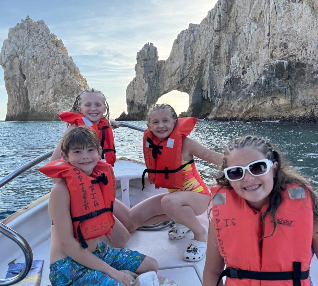 Four children wearing life jackets, smiling on a boat with the iconic Cabo Arch in the background during a sunny day on the water.