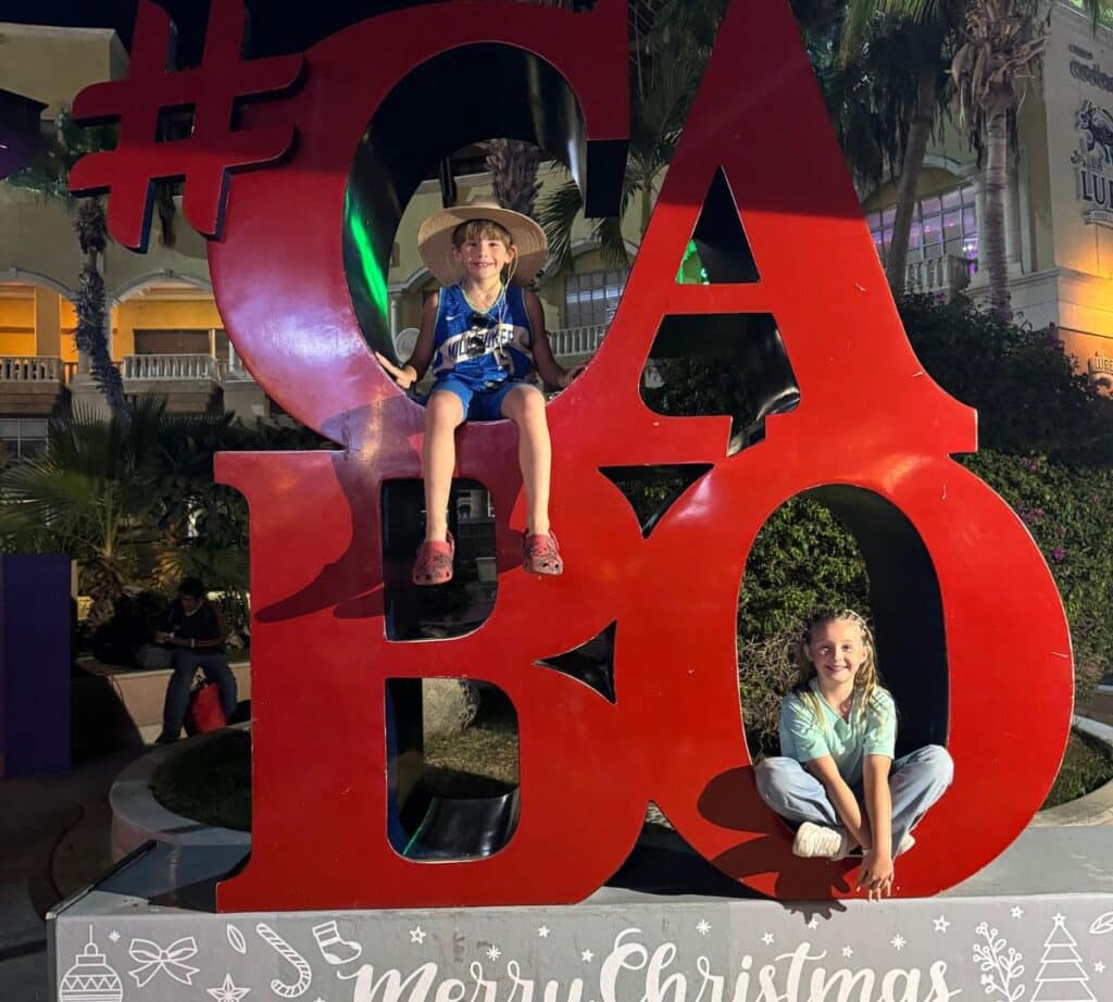 Children sitting on a festive "Cabo" sign, decorated with Merry Christmas and Happy New Year greetings, capturing the holiday spirit in downtown Cabo San Lucas.