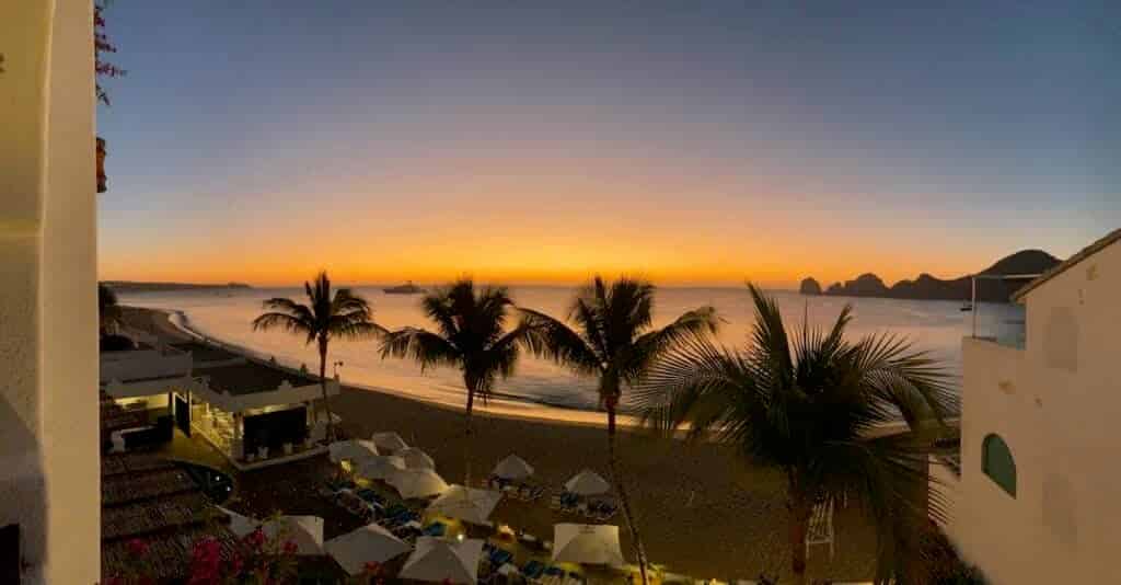 A stunning sunset view from the balcony of Pueblo Bonito Los Cabos, overlooking palm trees, the beach, and the ocean, with warm golden hues in the sky.