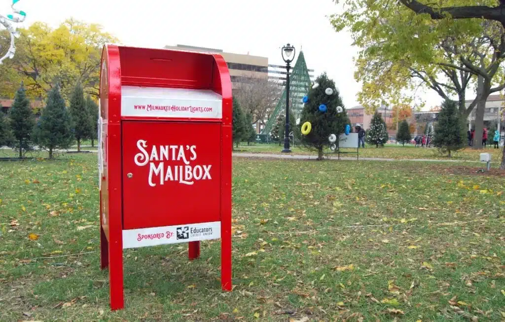 Red Santa's Mailbox in the middle of a town square and behind it are trees decorated with Christmas themes.