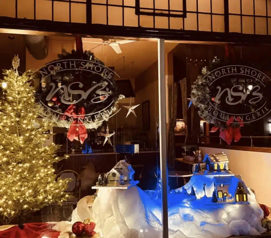 North Shore Boulangerie window decorated for Christmas with glowing wreaths, a lit tree, and a snowy village display.