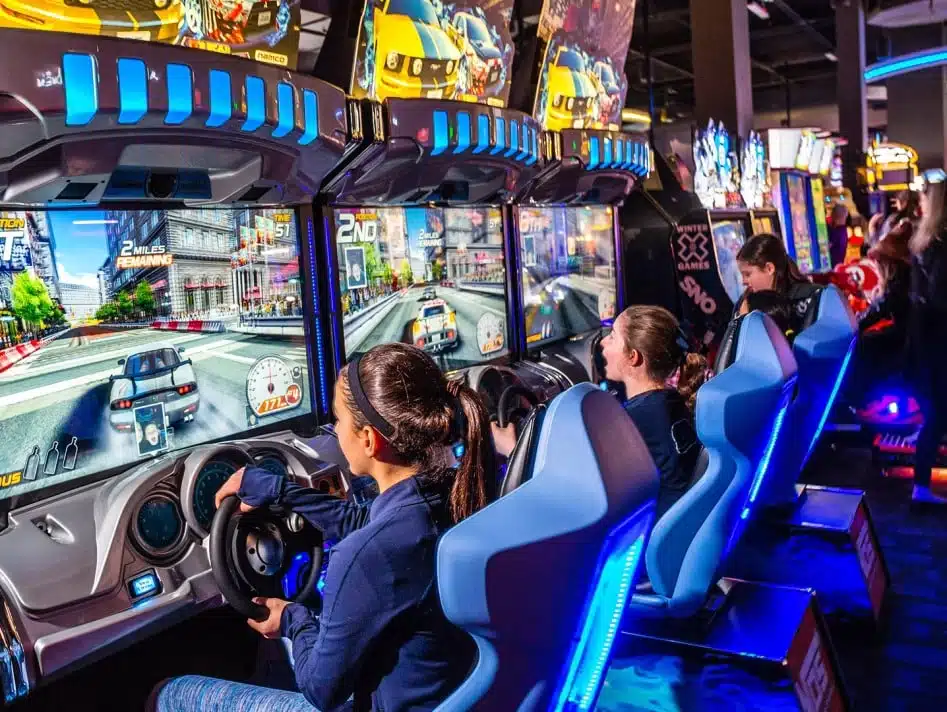 Kids enjoy a thrilling racing game at an arcade, steering through virtual city streets on brightly lit driving simulators.