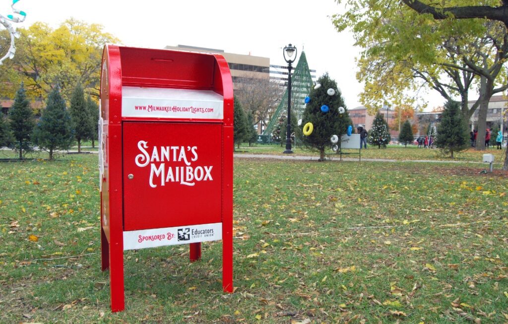 A red Santa's mailbox in the middle of a park and a Christmas tree in the background