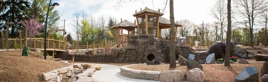 Playground featuring a large wooden treehouse structure, rock formations, climbing areas, and nature-themed play elements surrounded by trees.