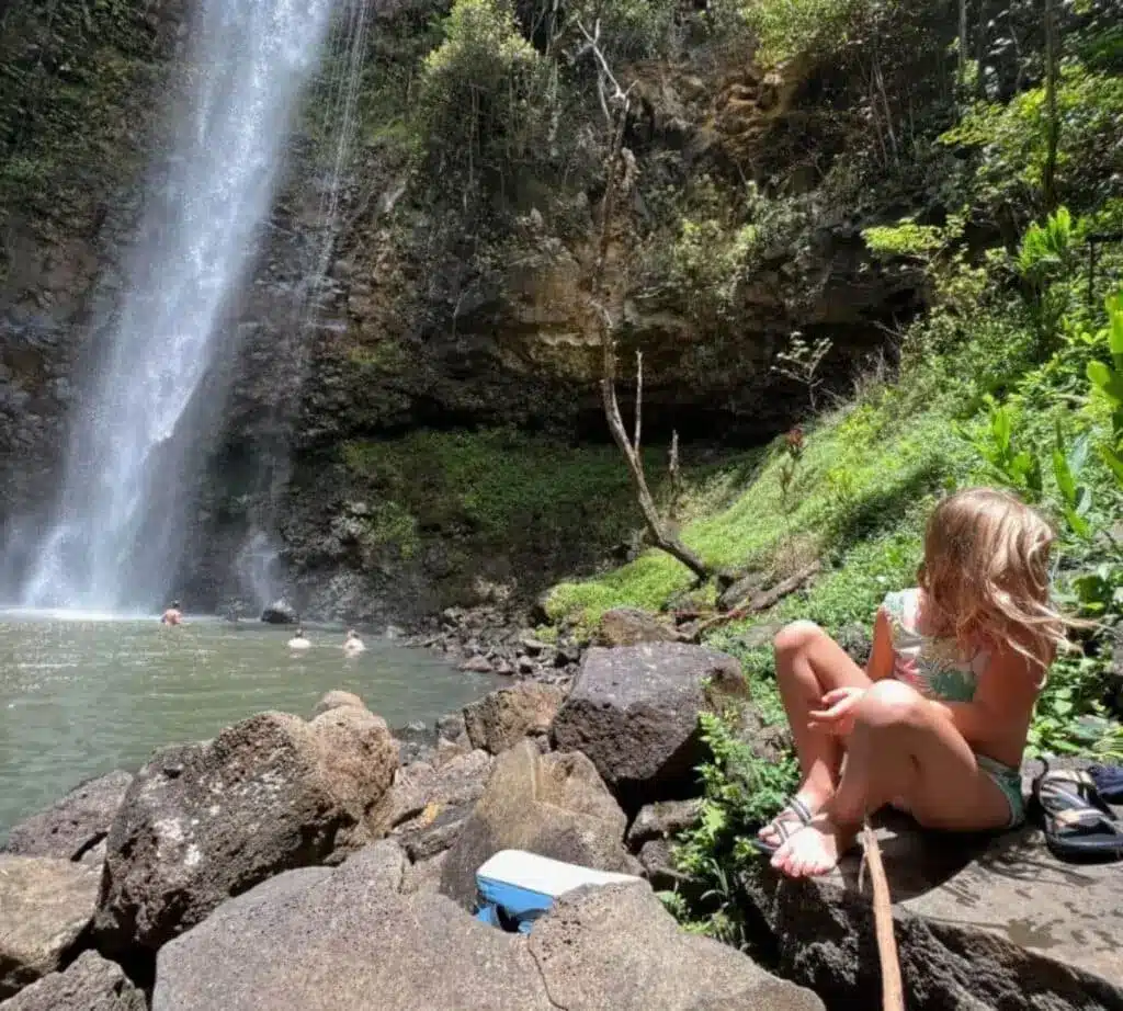 A child sitting on the rocks with a large falls in the background in Kauai