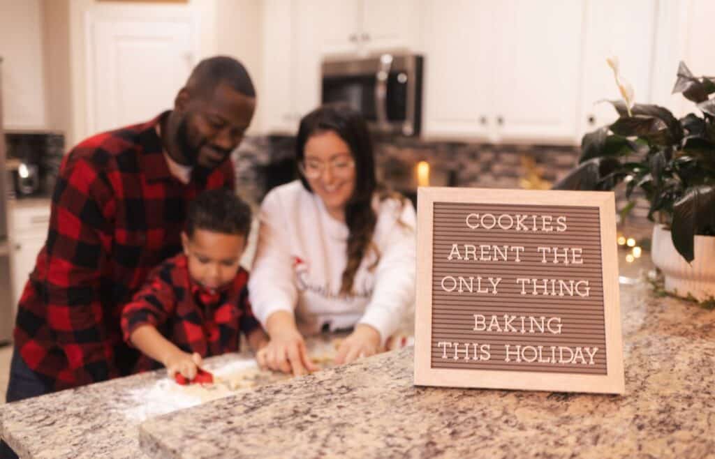 A family of three baking a cookie in the background with a letterboard that says Cookies arent the only thing baking this holiday in the foreground