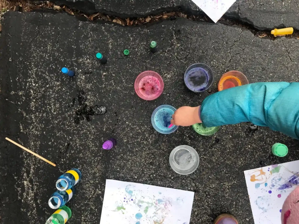 Child dipping straw into colorful bubble paint cups during outdoor bubble art activity.
