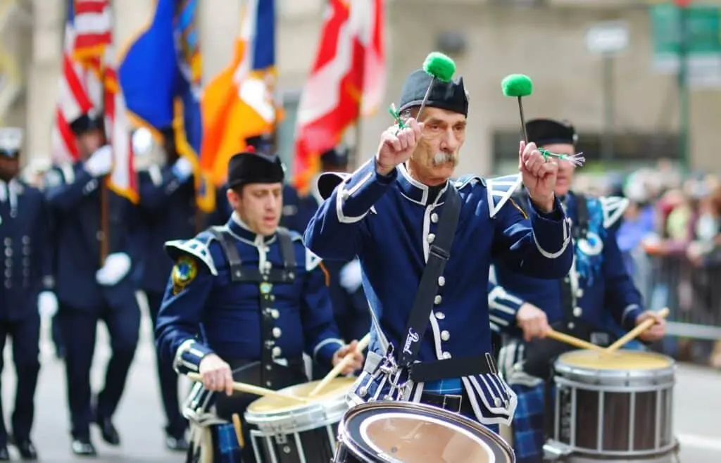 A marching band playing with the drummer holding green drum sticks with people 