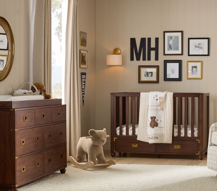 Neutral-toned baby nursery with wood furniture, including a crib and dresser, beige curtains, and framed wall art. A teddy bear quilt with the name 'Mason' hangs on the crib, a plush rocking bear sits on the rug, and a wall monogram with the initials 'MH' is displayed above the crib.
