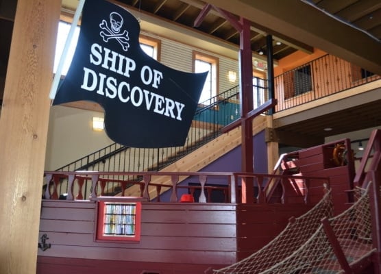 A pirate ship exhibit with a black flag and a view of wooden staircase with steel railing at Children's Play Gallery.