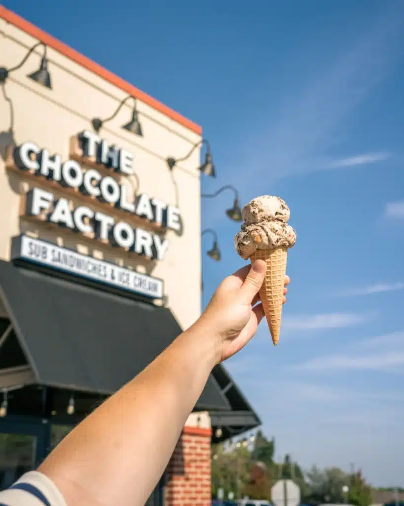 A hand holding a Rocky Road ice cream on a cone in front of the Chocolate Factory in Cedarburg