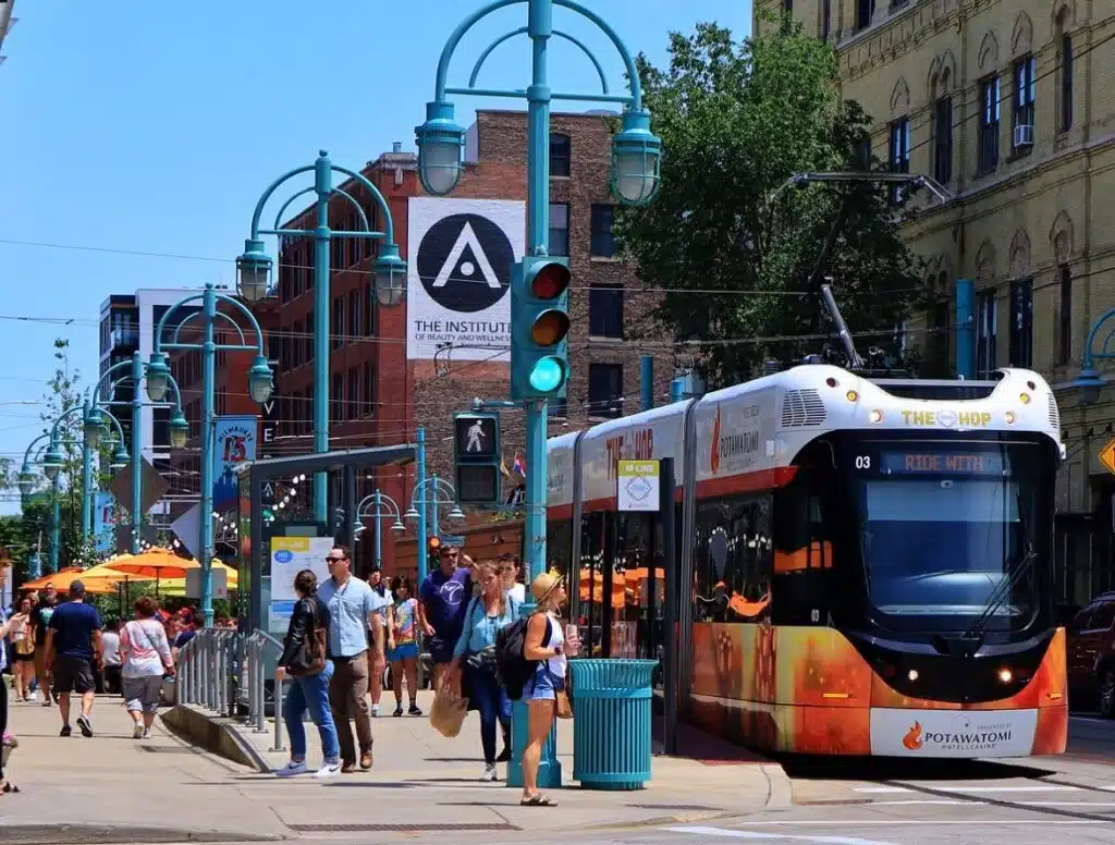 The Hop streetcar in downtown Milwaukee with pedestrians, city buildings, and outdoor dining on a sunny day.