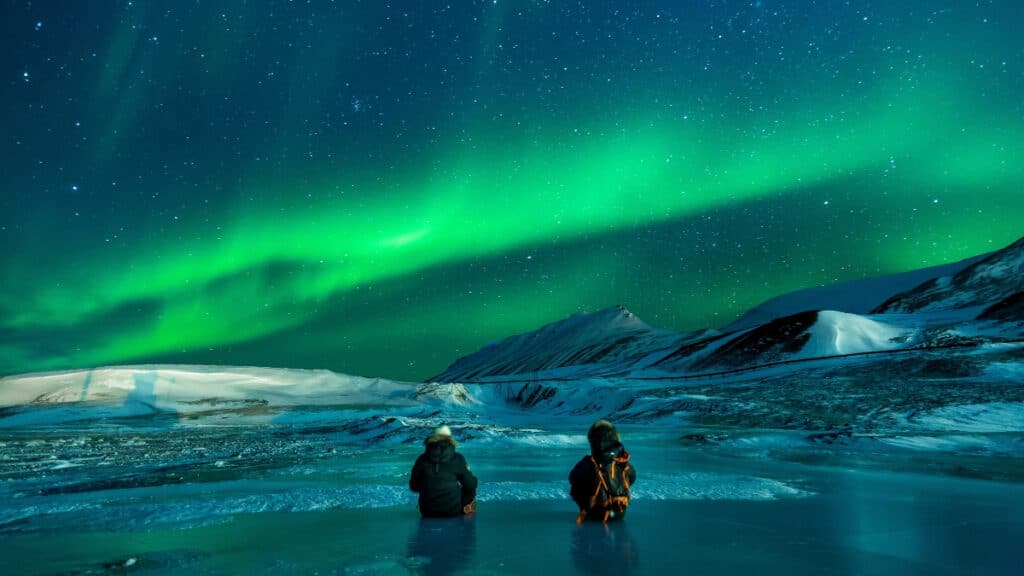 Two people stand in a glacial lake observing the green northern lights in the sky.