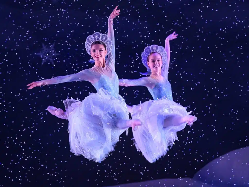 Two ballerinas with lace headdresses and silver blue costumes performing a grand jeté amidst the snow.