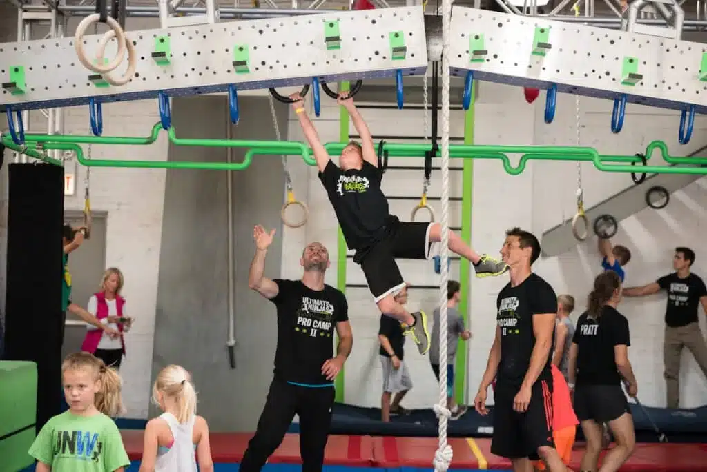A child hangs from overhead ninja obstacles while coaches supervise at Ultimate Ninjas Oak Creek.