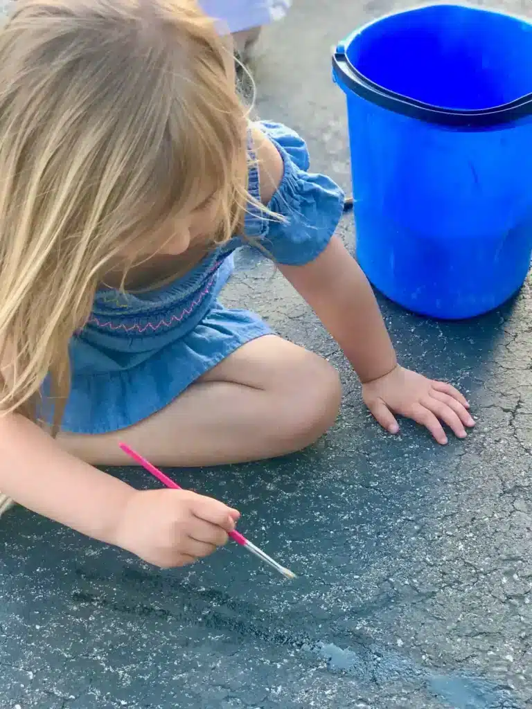 Child painting with water on pavement beside a bright blue bucket.