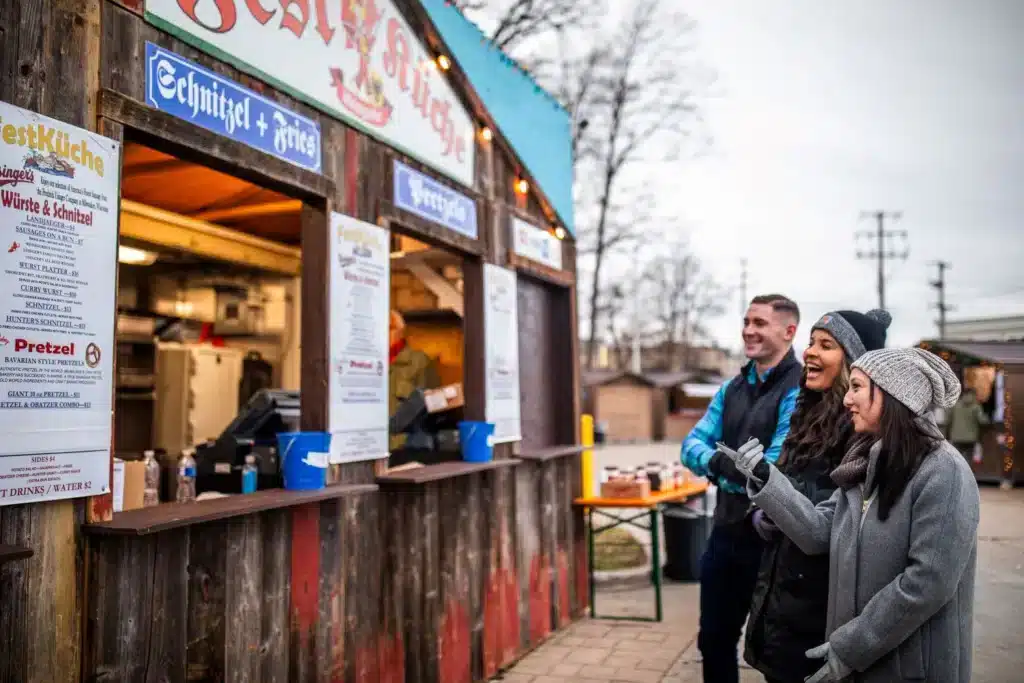 A Christmas market stall selling schnitzels, fries and pretzels with three smiling friends looking at the menu.