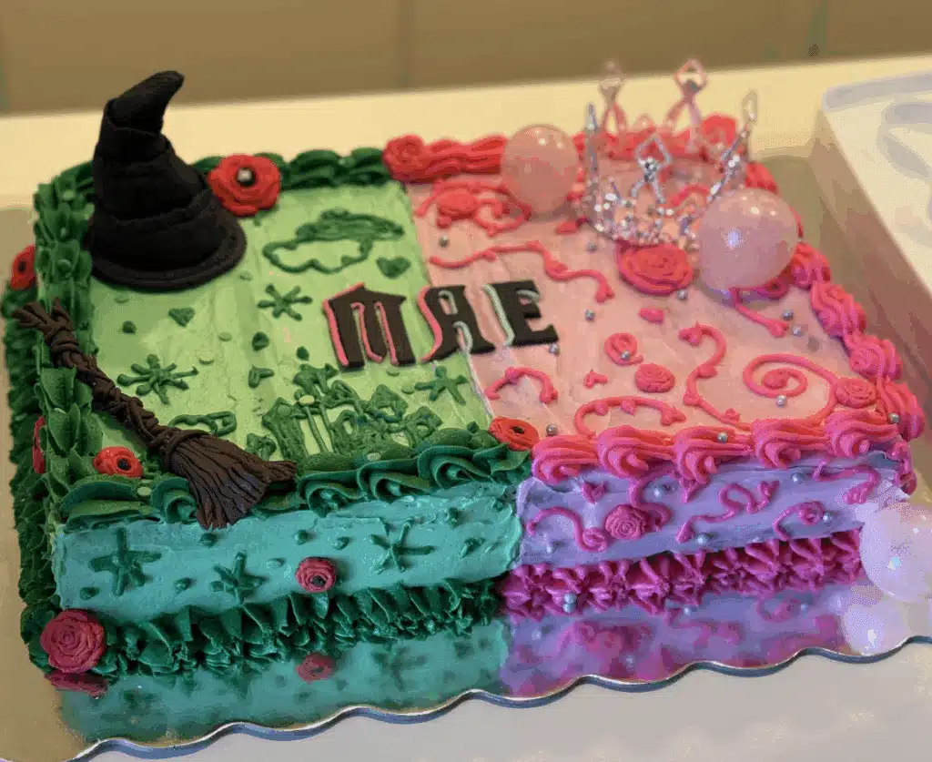 Wicked themed cake, one half green with a witch's hat and broom, and the other half pink with balloon bubbles and a silver crown.