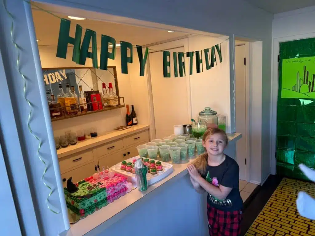Child stands by Wicked-themed party decorated, cake, and snacks, with a yellow brick road and poster of Oz in the background.