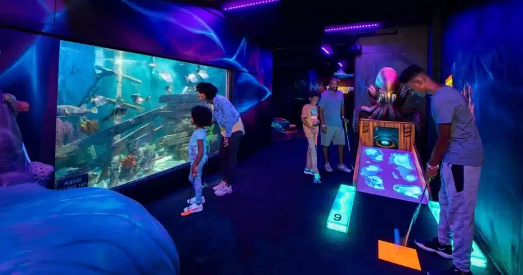 A family plays glow-in-the-dark, ocean-themed mini golf with tropical aquariums and underwater décor 