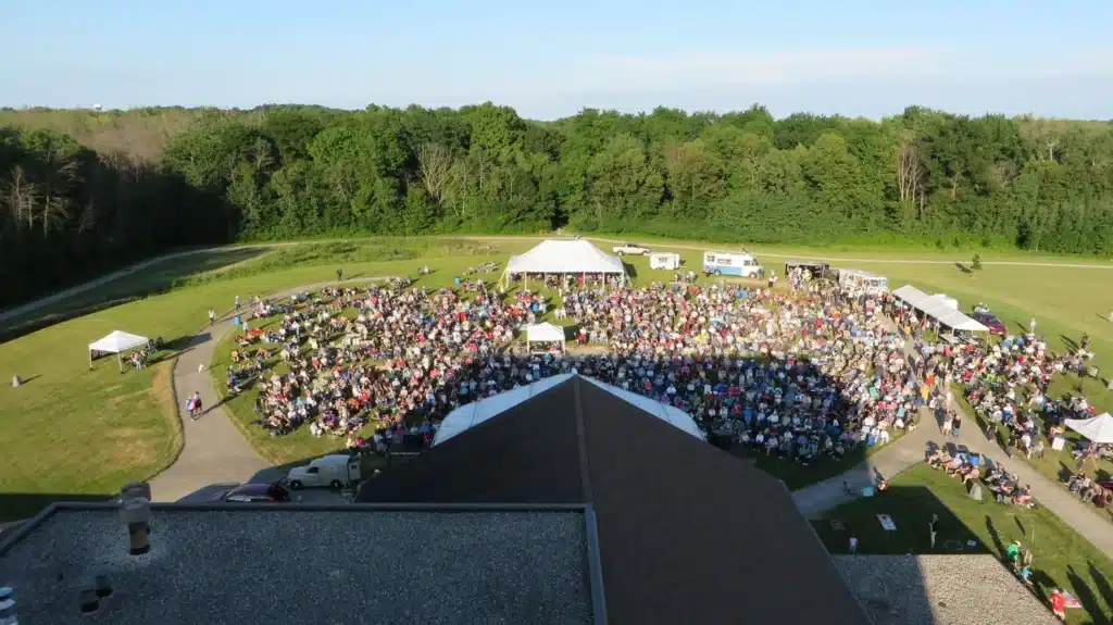 A large crowd gathers for an outdoor concert at the Sharon Lynne Wilson Center, with white tents, food trucks, and wooded surroundings.
