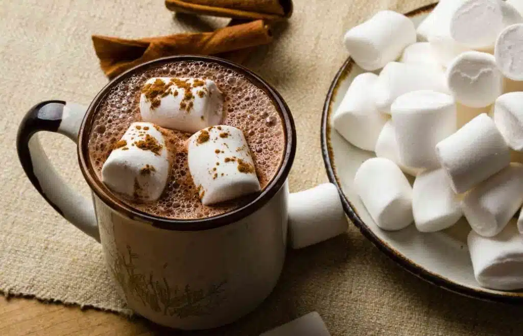 A mug full of hot cocoa with three large marshmallow and beside it a bowl of white marshmallows