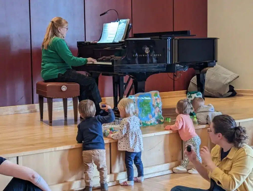 Toddlers play near piano while teacher performs during Smart Start early music class.